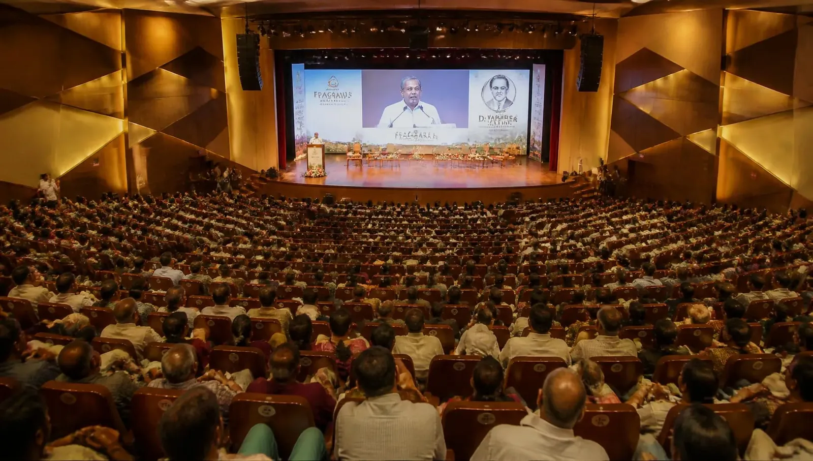 Packed auditorium during a live event seen from the rear balcony, speaker on stage